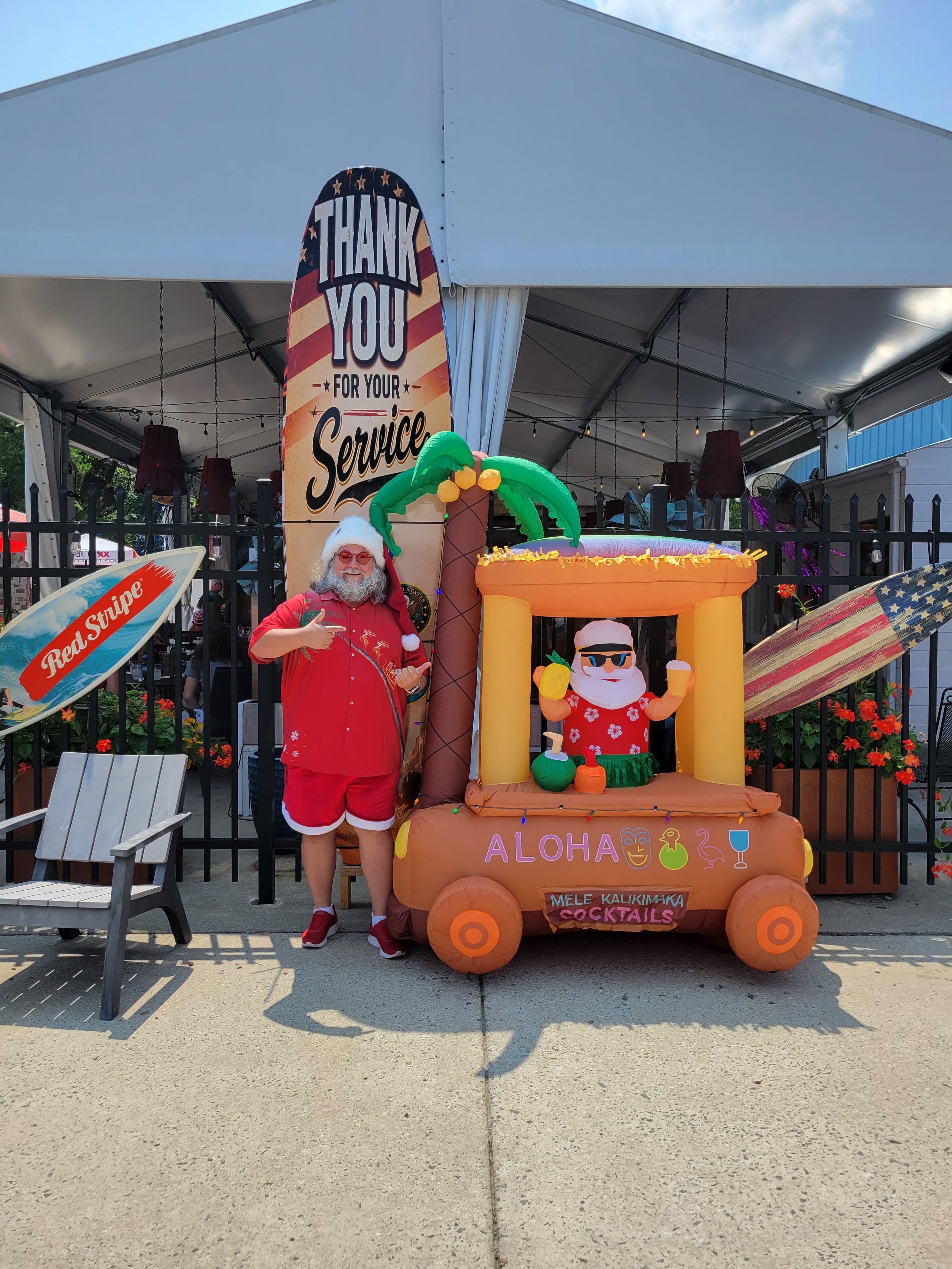 Cape May County Santa pointing at an Aloha Mele Kalikimaka Cocktails inflatable at an outdoor venue with a Thank You For Your Service surfboard