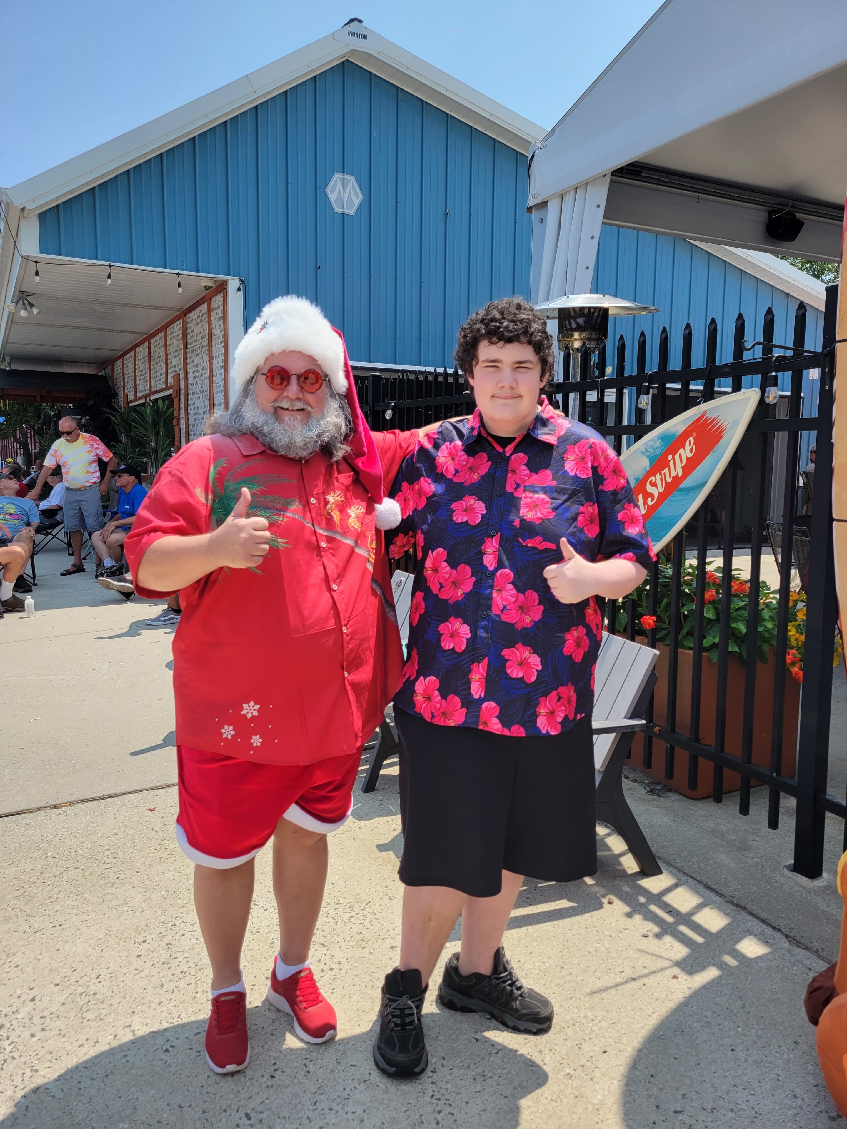 Cape May County Santa giving thumbs up with a teen in a pink floral Hawaiian shirt at an outdoor bar patio