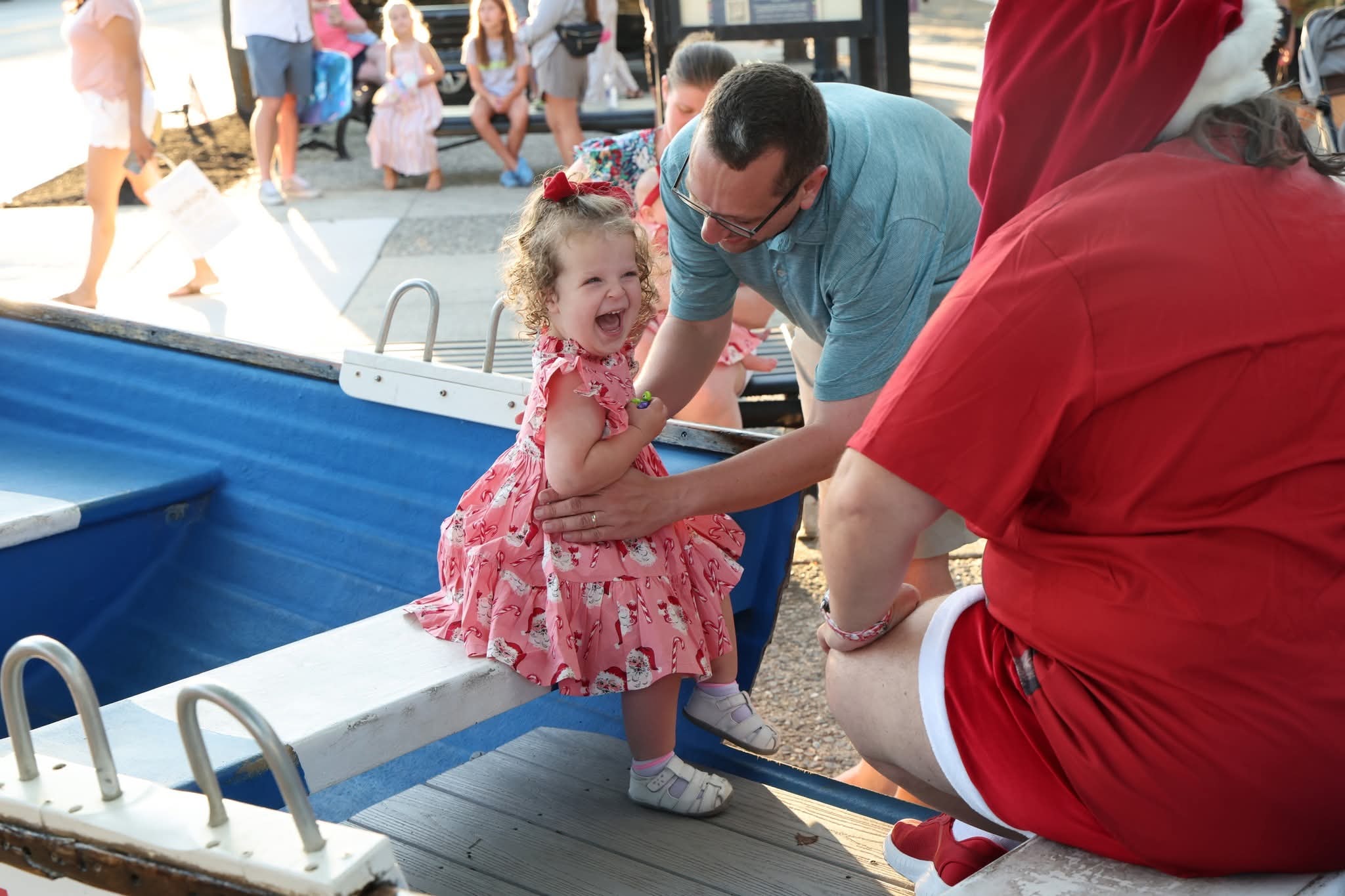 A toddler girl in a Santa-print dress laughing with pure joy as her dad helps her into the Stone Harbor boat